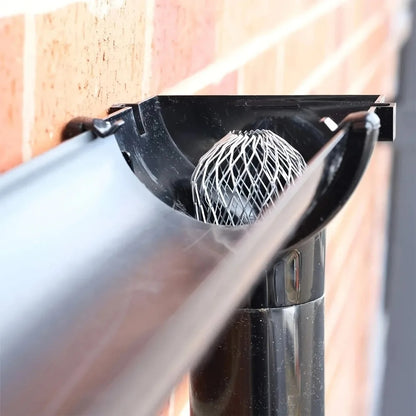 Metal mesh leaf strainer fitted inside a gutter outlet at the top of a downspout beside a brick wall.
